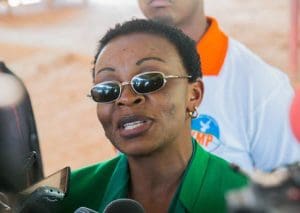 A closeup of Victoire Ingabire, wearing dark gold-rimmed sunglasses, a short haircut, and a collared shirt, speaking at a press conference.