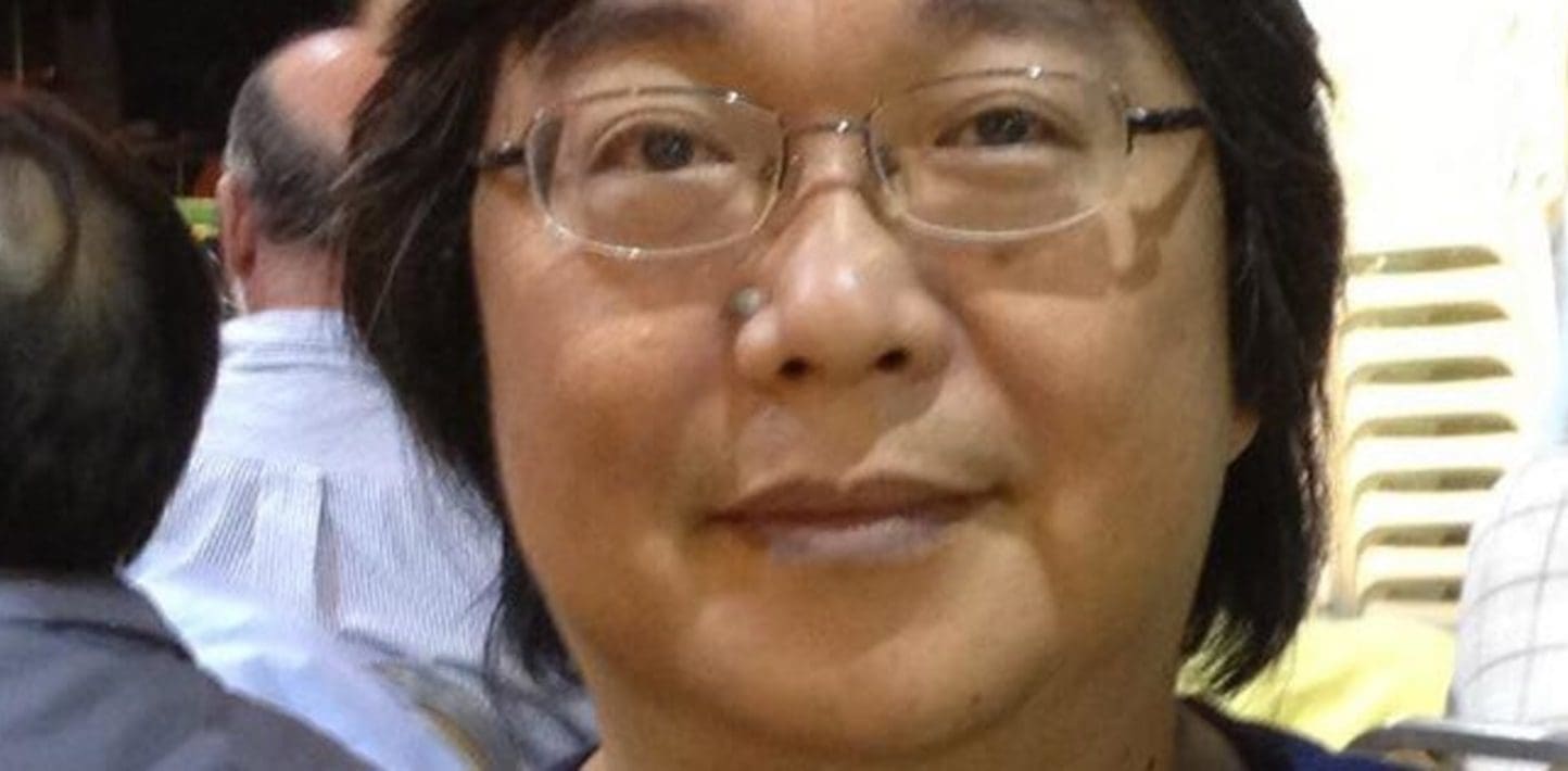 A blurry photo of Gui Minhai, his hair loose below his ears, wearing wireless glasses and looking into the camera in a crowded room.