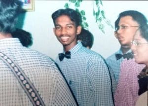 Pannir Selvam Pranthaman, wearing a checkered shirt with a bow tie, smiles at the camera surrounded by other young people wearing the same outfit, at an event with decorations on the walls. He has a thin moustache and parted hair.
