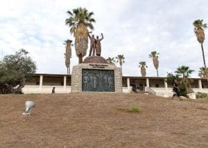 A stone and metal monument to the victims of Germany's genocide in Namibia stands on a a grassy hillside in front of a low building with palm trees around it. The monument has a stone frame, with an image made of metal inside of it depicting armed German soldiers standing around a tree with indigenous Africans hanging from the branches. On top of the stone frame is a metal sculpture of two figures with their fists raised in the air, with the words, 
