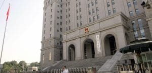 A tall, gray office building with an official Chinese government emblem consisting of a red circle with gold stars. The building has a long staircase leading up to arched entranceways. A Chinese flag hangs from a flagpole in front of the building. A security guard stands under an umbrella and a man in a white shirt speaks on a cellphone.