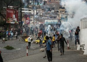 Tear gas clouds fill the air above as people run away on a crowded street strewn with bricks and burning wood.