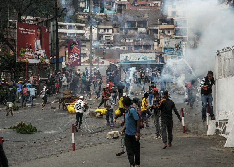 Tear gas clouds fill the air above as people run away on a crowded street strewn with bricks and burning wood.