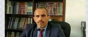 Abdul Majid Sabra, a middle aged man with short graying hair and a moustache, poses for a picture wearing a dark suit and red tie, seated at a desk in front of a dark bookshelf with many thick bound volumes.