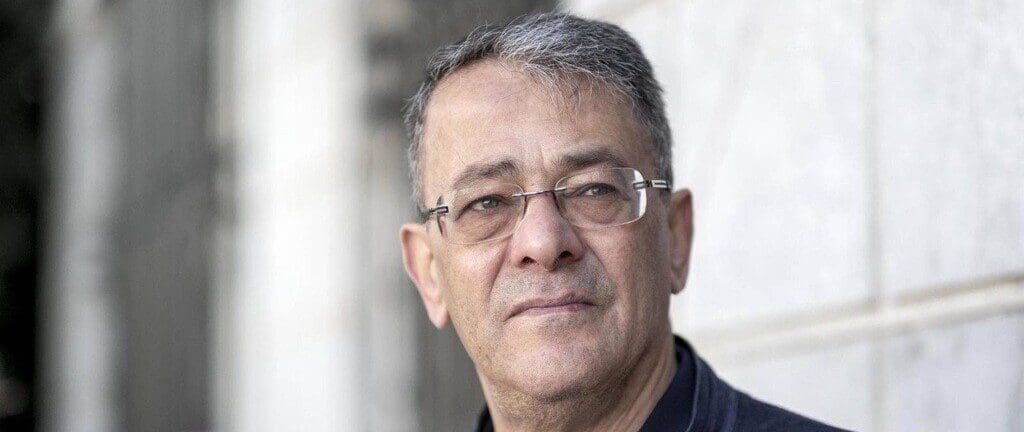 Ahmed Souab, with gray hair, loose strands poking above his forehead, wearing wireless glasses and what appears to be a dark jean jacket over a dark sweater looks off to his left, with light falling on the right side of his face.