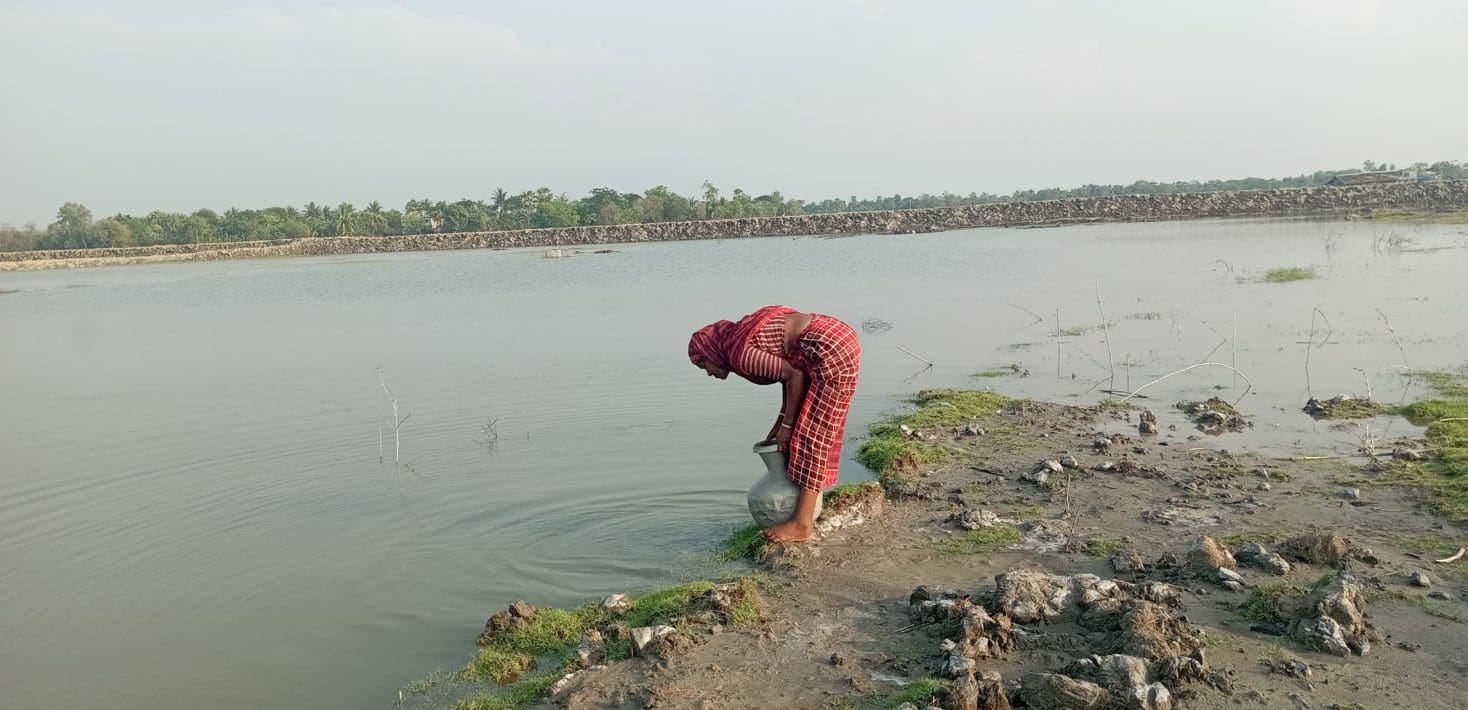 A young woman in Bangladesh bends over a river, holding a silver basin.