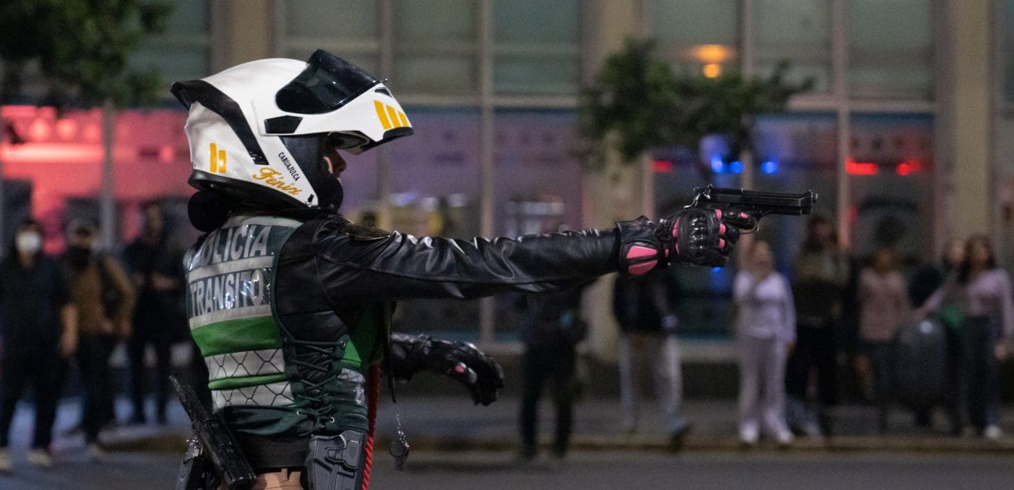 Person in a helmet, leather jacket, vest that says Policia holding pistol up