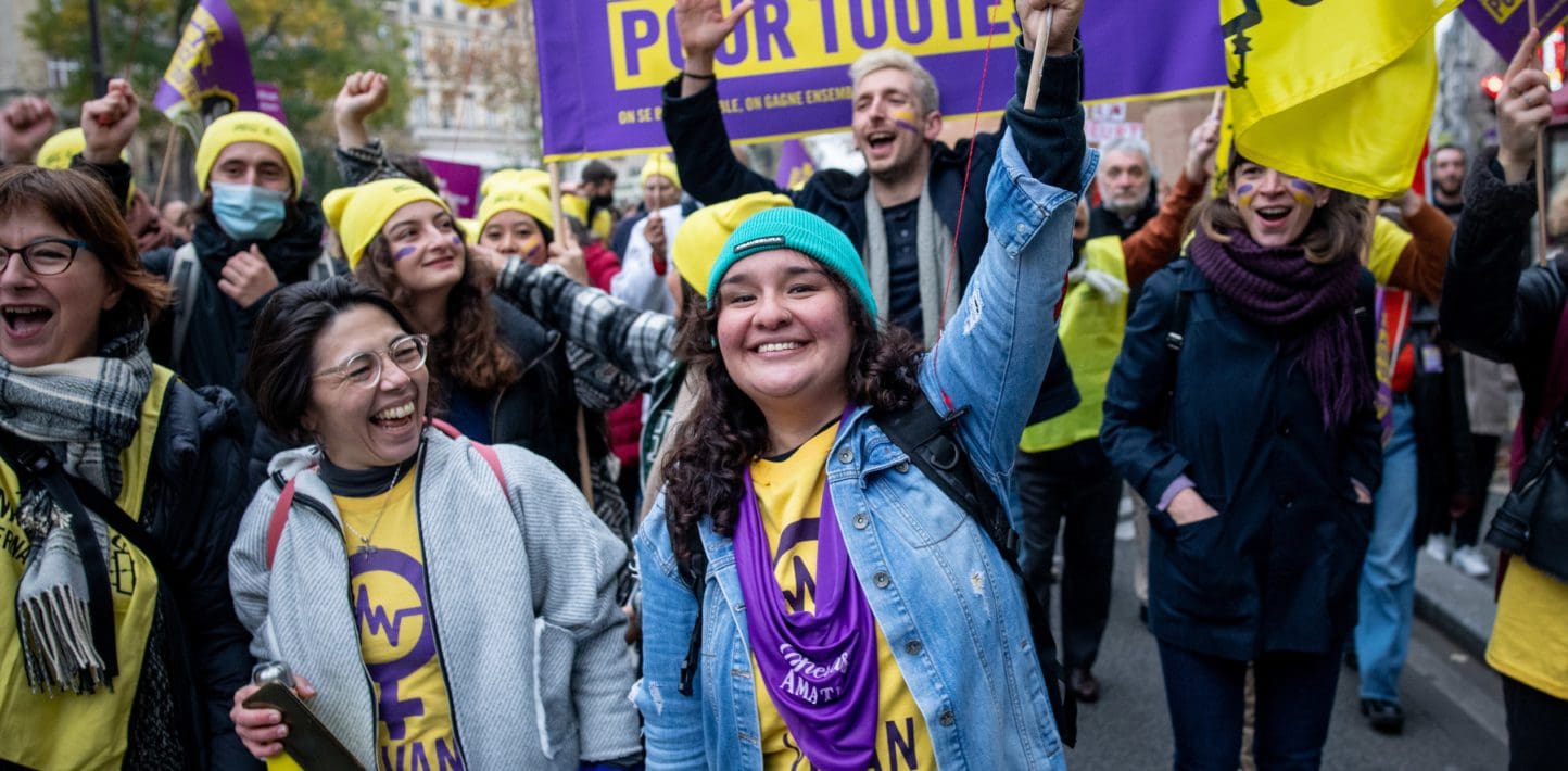 A woman smiling at the camera, surrounded by a group of people cheering at a protest in France. Everyone is wearing purple and yellow clothing.