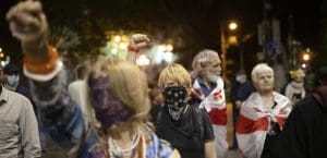 Multiple people attending a protest in Georgia, holding their fists in the air. The photo is focused on the center, where a young woman wearing all black holds her right fist in the air, surrounded by others at the protest.