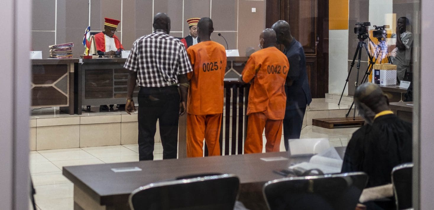 Two men wearing orange jumpsuits stand in a courtroom before two judges.