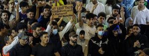 A crowd of young men, many shouting or holding their fists in the air. One holds up a cardboard sign that reads: 