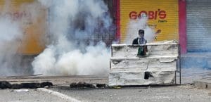 A man in a black cap and black shirt with a black and white scarf wrapped around his face stands behind a torn metal barricade in front of a cloud of tear gas on an otherwise deserted street.