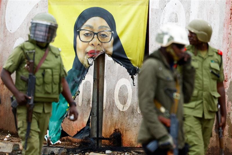 Armed police with green uniforms, magazine pouches, helmets, and AK-47 rifles walk past a ripped campaign poster of Tanzania's President Samia Suluhu Hassan.