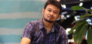 Y Quynh Bdap, a young man, sits next to a green leafy plant in front of painted wall, looks at the camera. He wears a blue and black plaid short sleeved collared button down shirt, which is open at the neck. He has a scruffy beard and thin moustache. His head is tilted to his left.