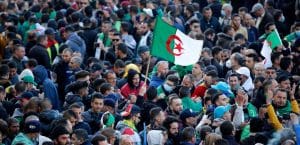A crowd of men, looking festive and joyous, wearing winter clothes. One holds an Algerian flag aloft on a thin pole, it whips in the wind. Some people take pictures of each other. The protest is joyous.