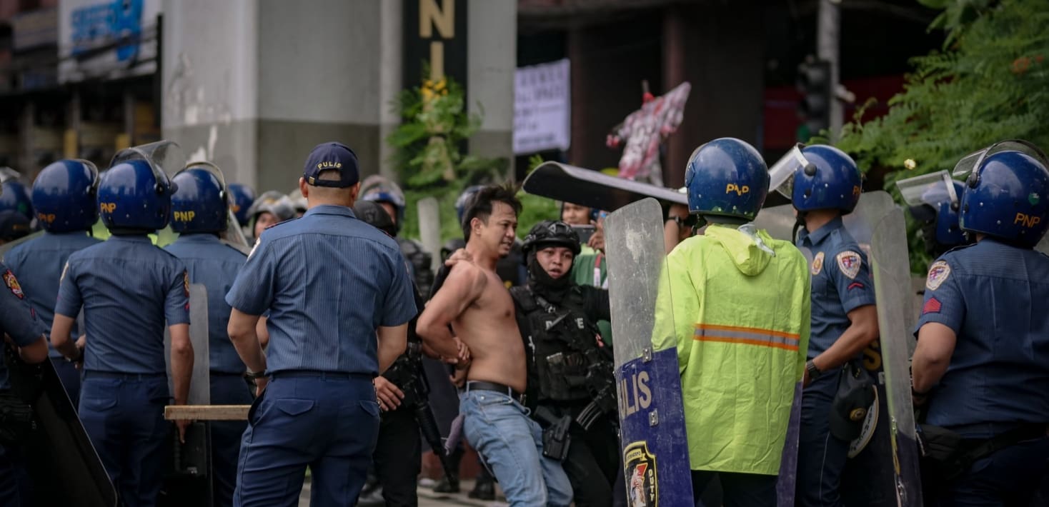 Police in riot gear push a shirtless man whose hands are cuffed behind his back.