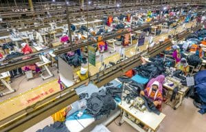 A bird's eye view of a factory warehouse with dozens of women stitching clothes with sewing machines.