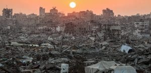 A skyline image of Gaza Strip, with destroyed buildings and debris, and a sunset in the background.