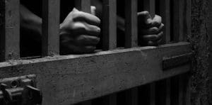 A black and white image of prison bars held by two hands.