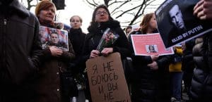 Protesters with somber faces wearing all black hold pictures of Aleksei Navalny, flowers, and signs.