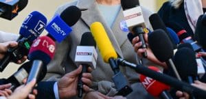 Dozens of news broadcast microphones aimed at a faceless speaker wearing a gray coat.
