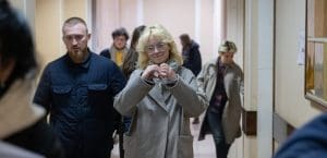 A blonde woman in handcuffs, holding her hands up in the shape of a heart.