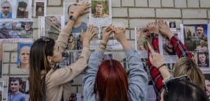 Three women taping posters of male Ukrainian POWs on a white wall.