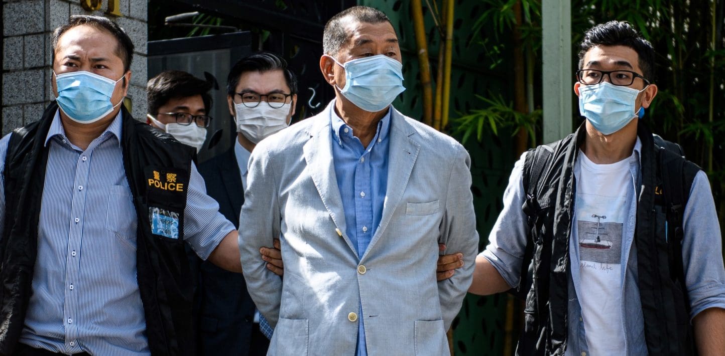 Jimmy Lai, wearing a gray linen suit, blue shirt, and a facemask, walks out of a darkened room into sunlight, his hands behind his back, held on each side by masked police officers. Lai's eyebrows are raised. His chin lifted up.