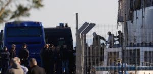 Two blue vans are parked outside of a building with barbed fence. A group of people stand outside of the building. One man is being led by two other men, from the building to the blue vans. He is in handcuffs.
