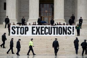 A group of protesters stands at the steps of the Supreme Court with a huge black and white banner reading STOP EXECUTIONS. Police in dark uniforms circle around the group.