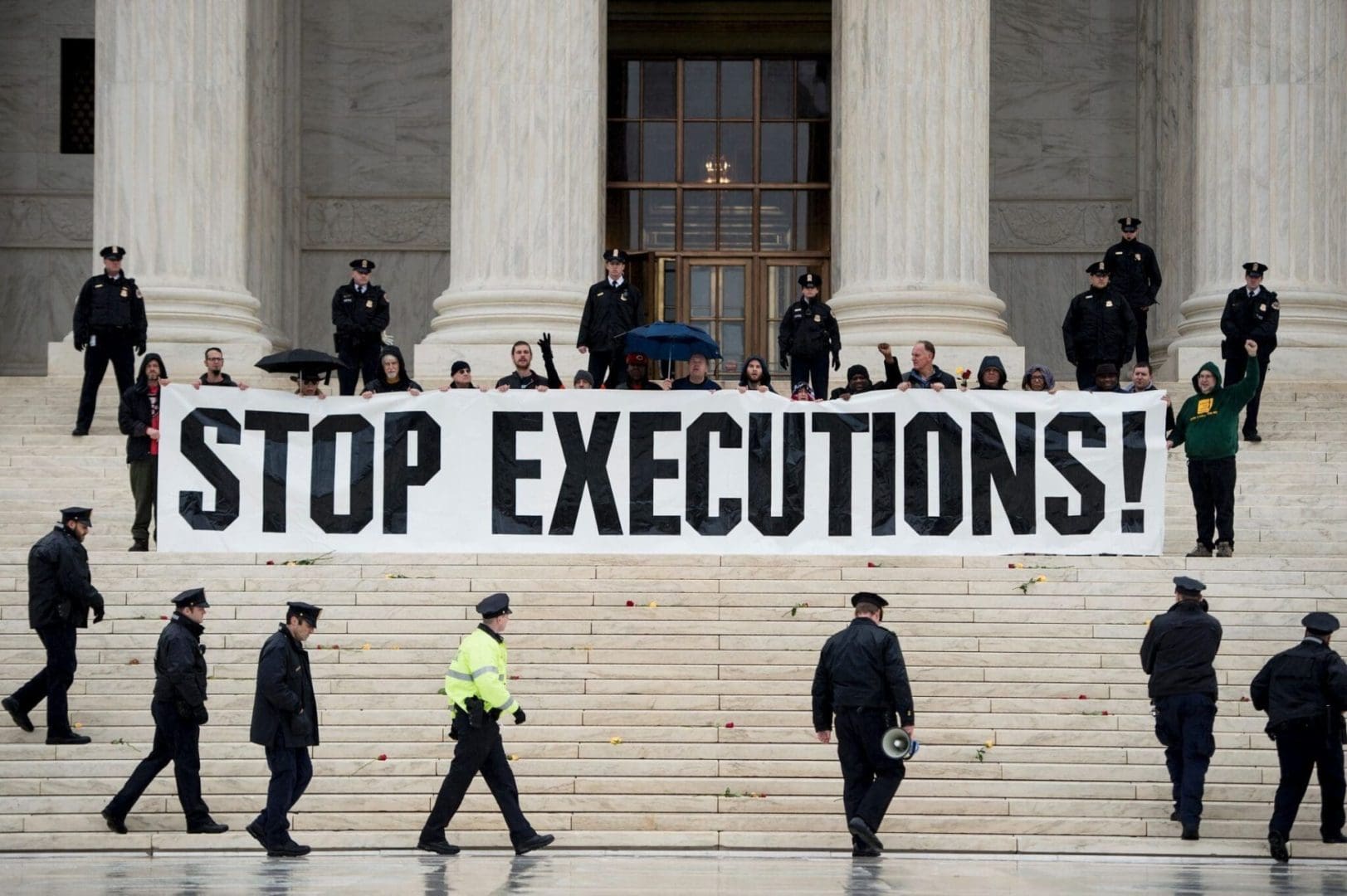 A group of protesters stands at the steps of the Supreme Court with a huge black and white banner reading STOP EXECUTIONS. Police in dark uniforms circle around the group.