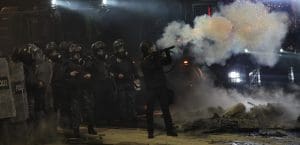 A group of riot police dressed in all black and equipped with shields. One police officer is holding a flame thrower.