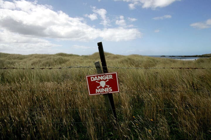 A green field surrounded by a wired fence. There is a red sign on the fence. The red sign says 