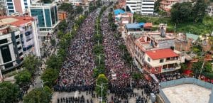 Streets filled with protesters stretching beyond the frame, with a thin line of police in riot gear blocking the front.