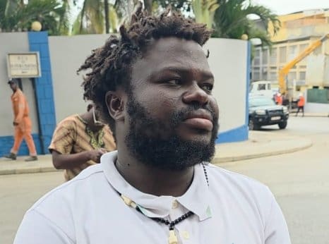 Serrote Jose de Oliveira, with a beard, stands in a street looking off to his left, his chin raised slightly. His shirt is white with a black necklace.