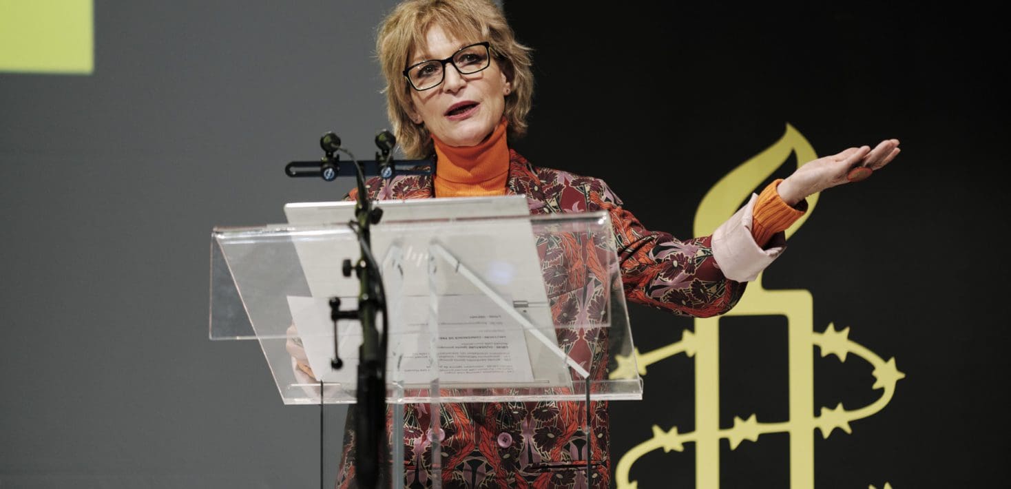A woman in black glasses and a colorful suit and turtleneck sweater speaks before a clear podium in front of Amnesty's black and yellow candle logo. She is gesturing with her left hand as she talks.