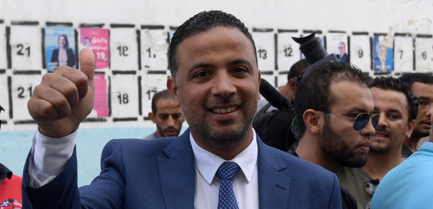 Seifeddine Makhlouf, wearing a blue suit and tie with a white shirt, holds a thumbs up and smiles in a crowded room. He has a stubbly gray beard and hair slicked back.