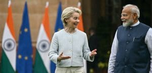 A photograph of Ursula von der Leyen, President of the EU Commission speaking with Narendra Modi, Prime Minister of India. Ursula is a middle-aged, white woman with short grey hair, wearing a button-down white blazer jacket. Modi is a middle-aged South Asian male wearing a black Indian Bandi vest. They are walking and talking in front of India and EU flags.