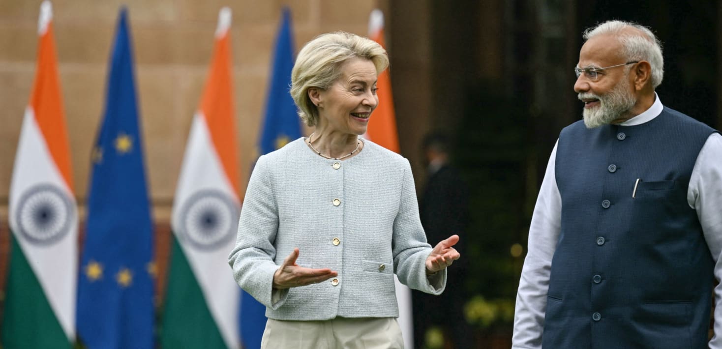A photograph of Ursula von der Leyen, President of the EU Commission speaking with Narendra Modi, Prime Minister of India. Ursula is a middle-aged, white woman with short grey hair, wearing a button-down white blazer jacket. Modi is a middle-aged South Asian male wearing a black Indian Bandi vest. They are walking and talking in front of India and EU flags.