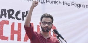 A young man speaks into a microphone with his fist raised above his head, a banner behind him.