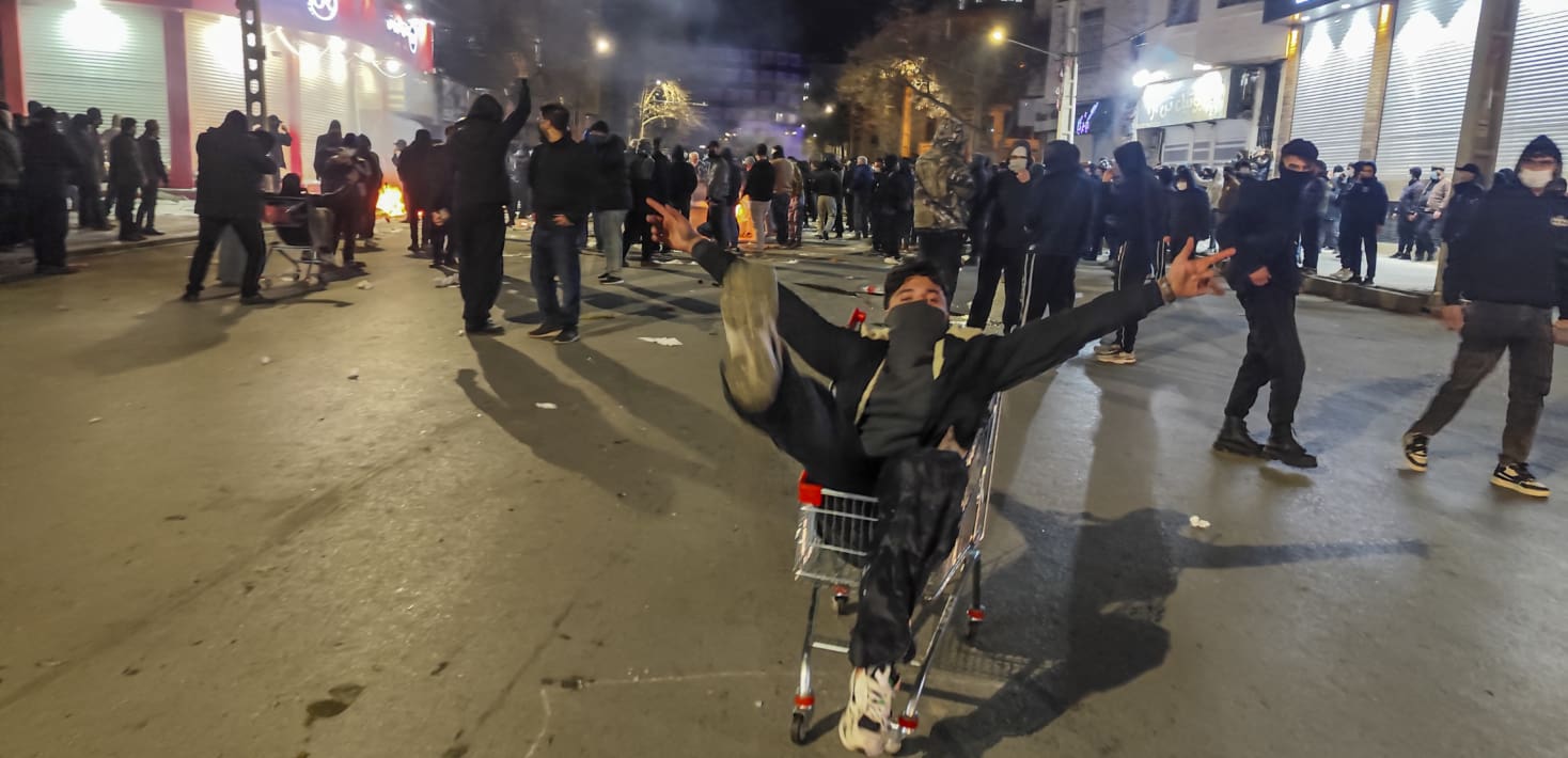 A young person rides a shopping cart and poses with a bandana around his face and his arms outstretched amid protesters filling a street, mostly wearing black. Many have their faces covered. There are small fires and smoke in the air.
