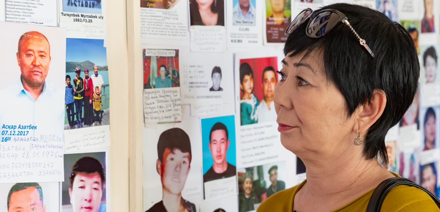 A middle aged Asian woman with short black hair, looking at a wall of photographs. The photographs are of ethnic Kazakh men who went missing in the Xinjiang Uyghur Autonomous Region.