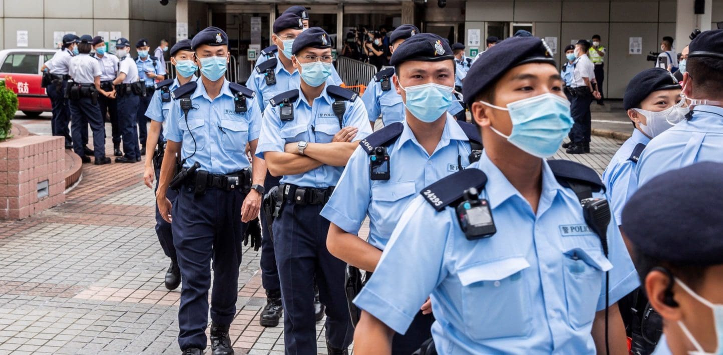 Rows of uniformed police wearing medical masks walk stand outside a building on a city street.