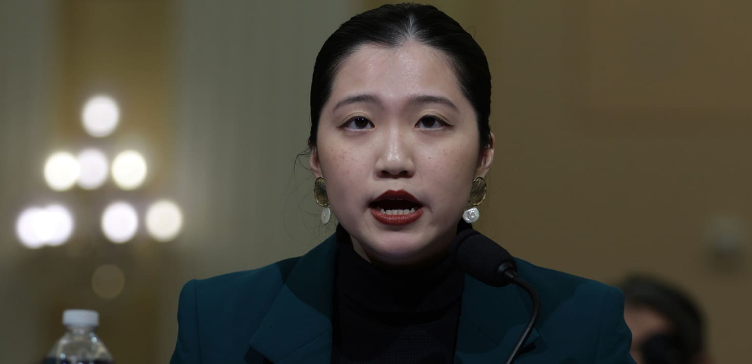 Anna Kwok, wearing a dark suit and turtleneck, faces forward and speaks in a room lit by dim lights behind her. She wears large earrings and her hair is pulled back and parted in the middle.