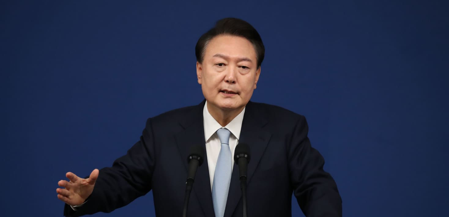 Yoon Suk Yeol, wearing a dark suit, white shirt, and light blue tie, speaks in front of two microphones, gesturing with his right hand, in front of a dark blue background.