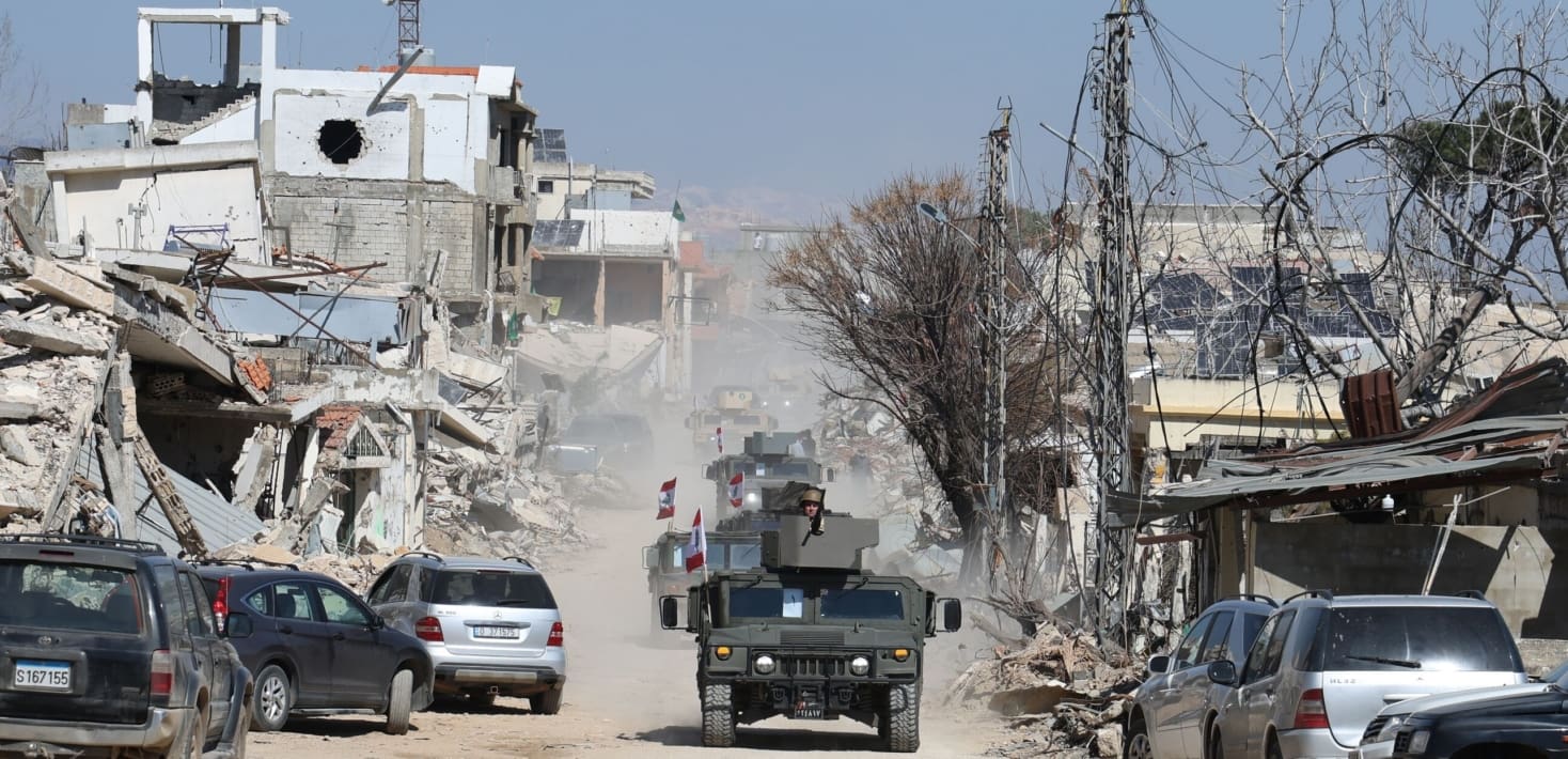 Black army car and other cars among dusty road with destroyed buildings