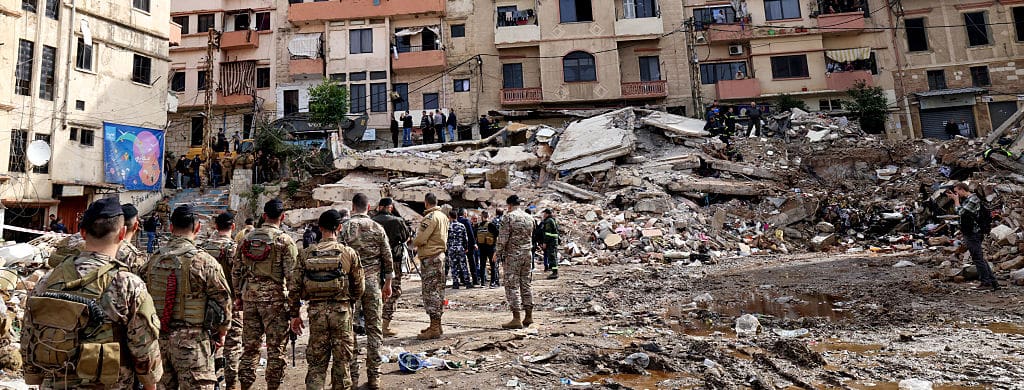 Soldiers in uniform stand amid a huge pile of rubble surrounded by apartment buildings in a dense residential neighborhood.