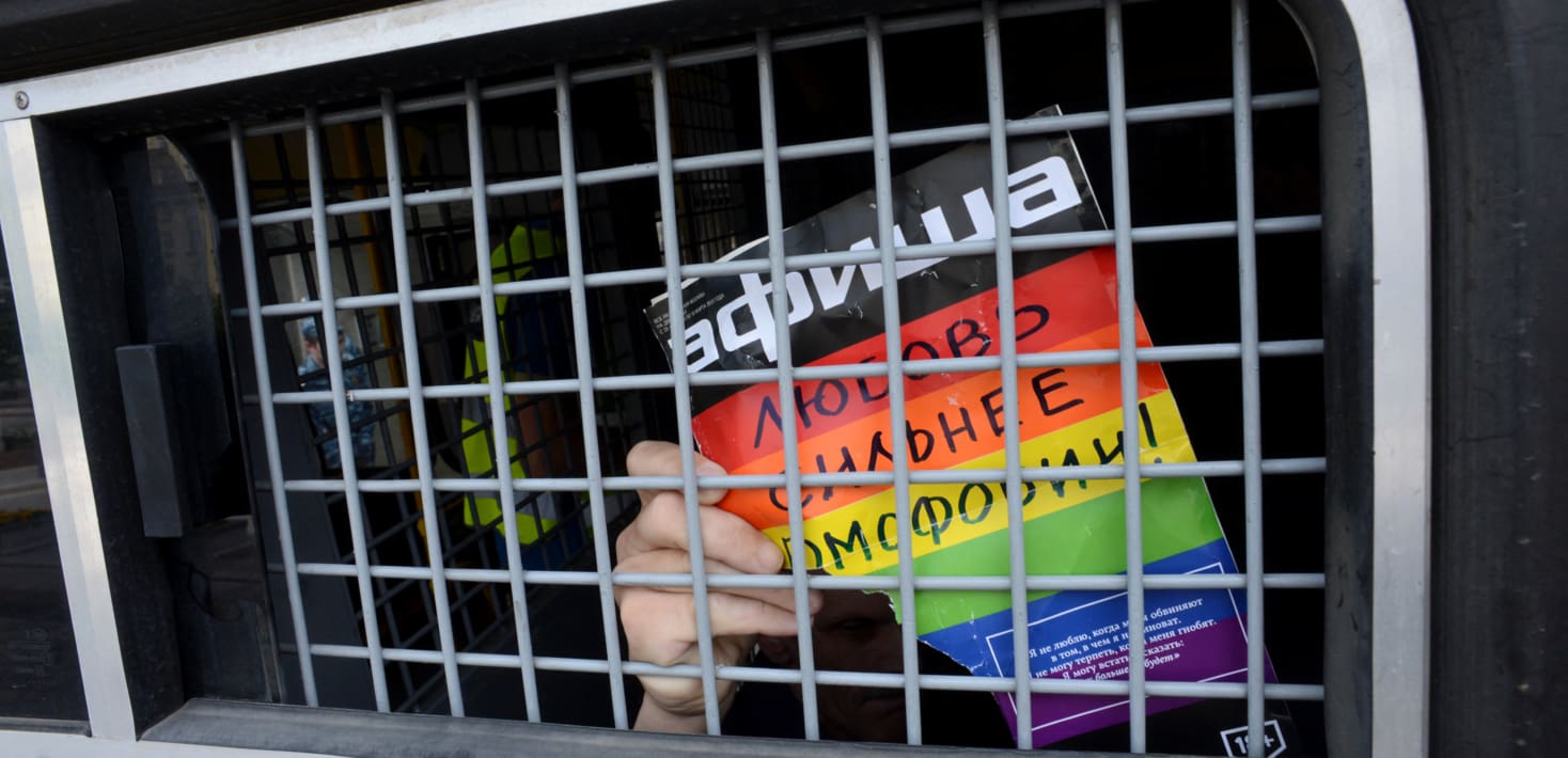 A person in the back of a police van, holding a rainbow-colored poster written in Russian.