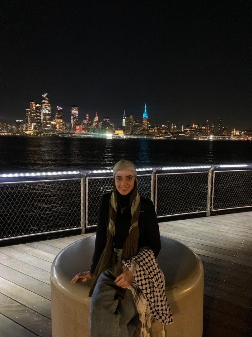 A smiling, young Palestinian woman wearing a hijab, black turtleneck, and jeans - while holding a Keffiyah. She is sitting on a pier in front of a city skyline at night.