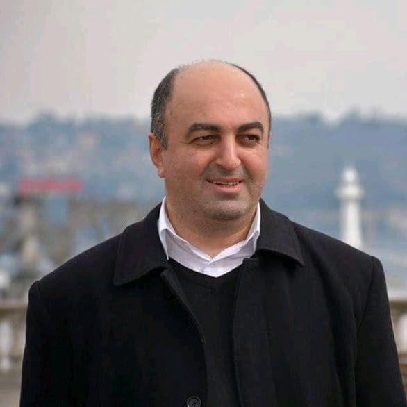 A smiling, bald, middle-aged Caucasian man wearing a black jacket, black sweater and white collared shirt.